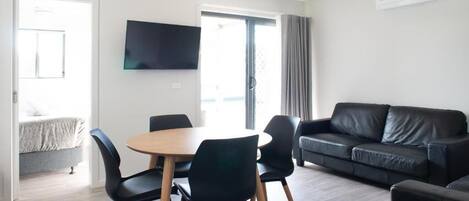 A modern living room with a round wooden table, black chairs, a black leather sofa, and a flat-screen TV mounted on the wall.