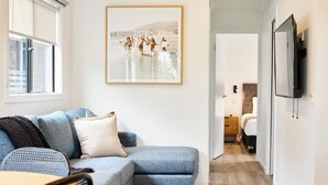 Een moderne woonkamer met een blauwe bank, een ingelijst schilderij aan de muur, en een aan de muur bevestigde televisie.