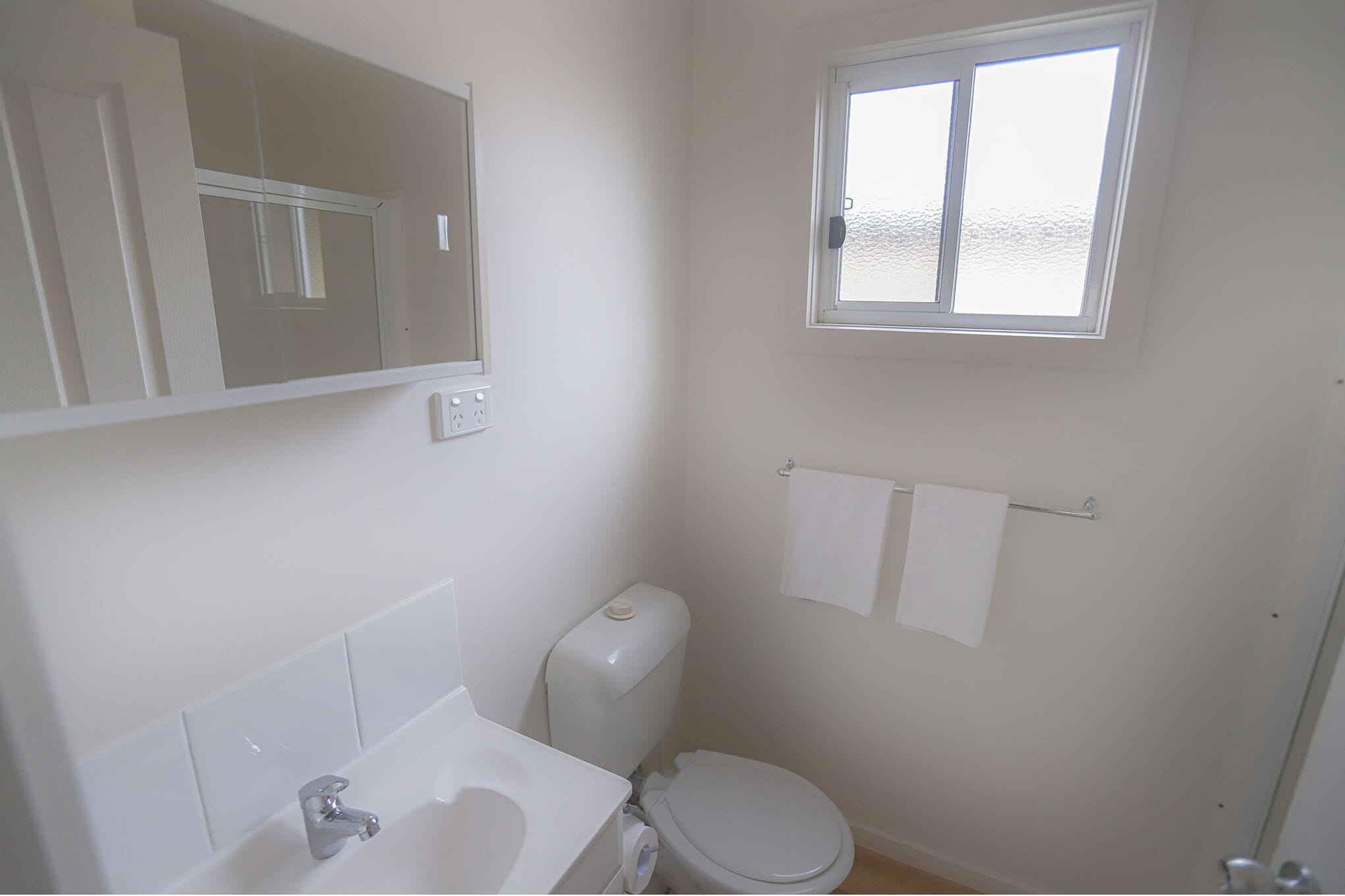 A small, clean bathroom with a toilet, sink, mirror, and window.