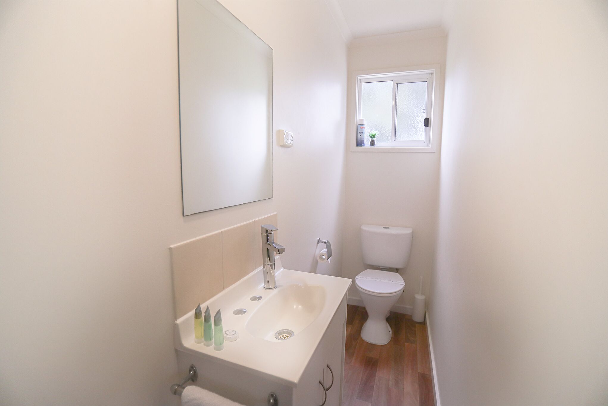 A small, white bathroom with a sink, mirror, toilet, and window.