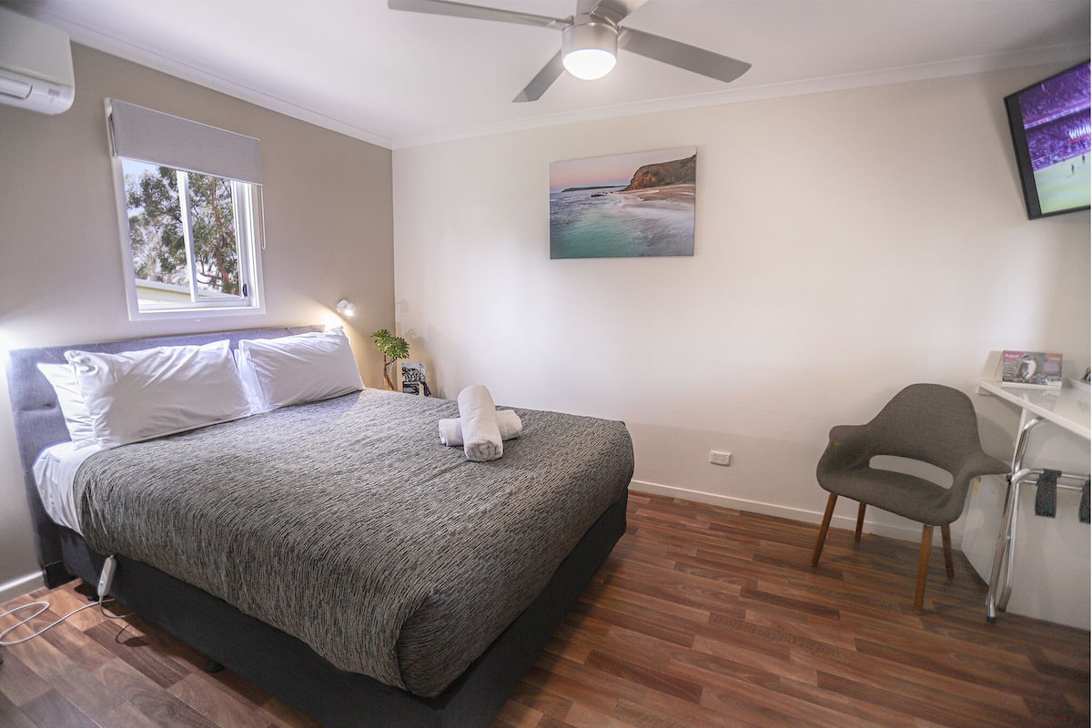 A bedroom with a bed, a ceiling fan, a window, a painting, and a desk with a chair.