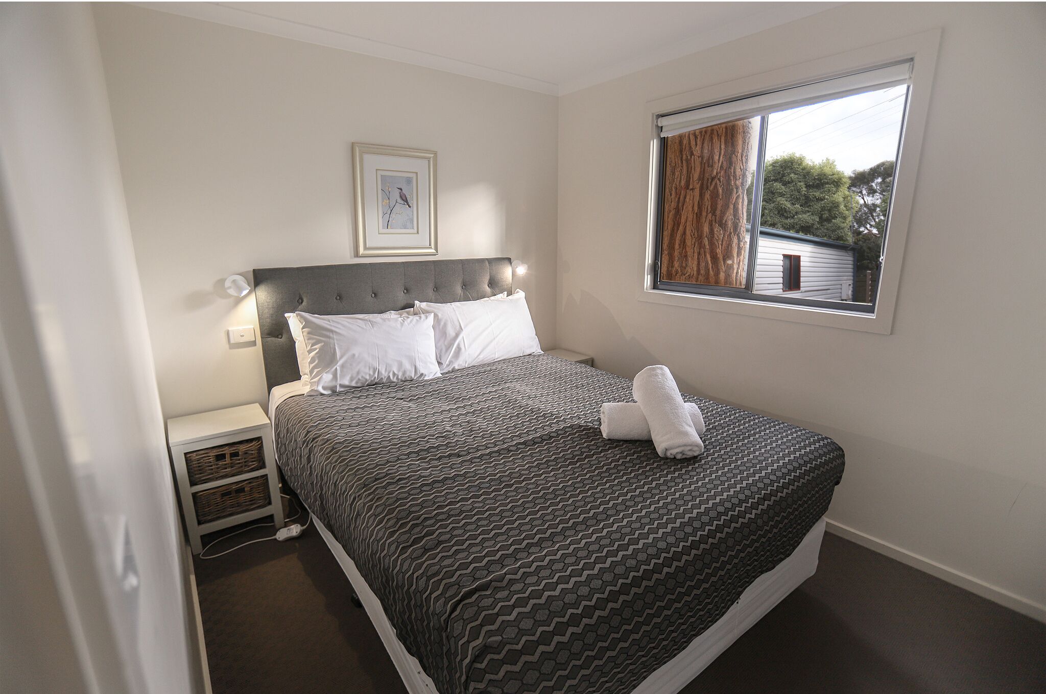 A neatly made bed with a grey patterned bedspread and white pillows, a bedside table with a lamp, and a window with a view of a house and trees.