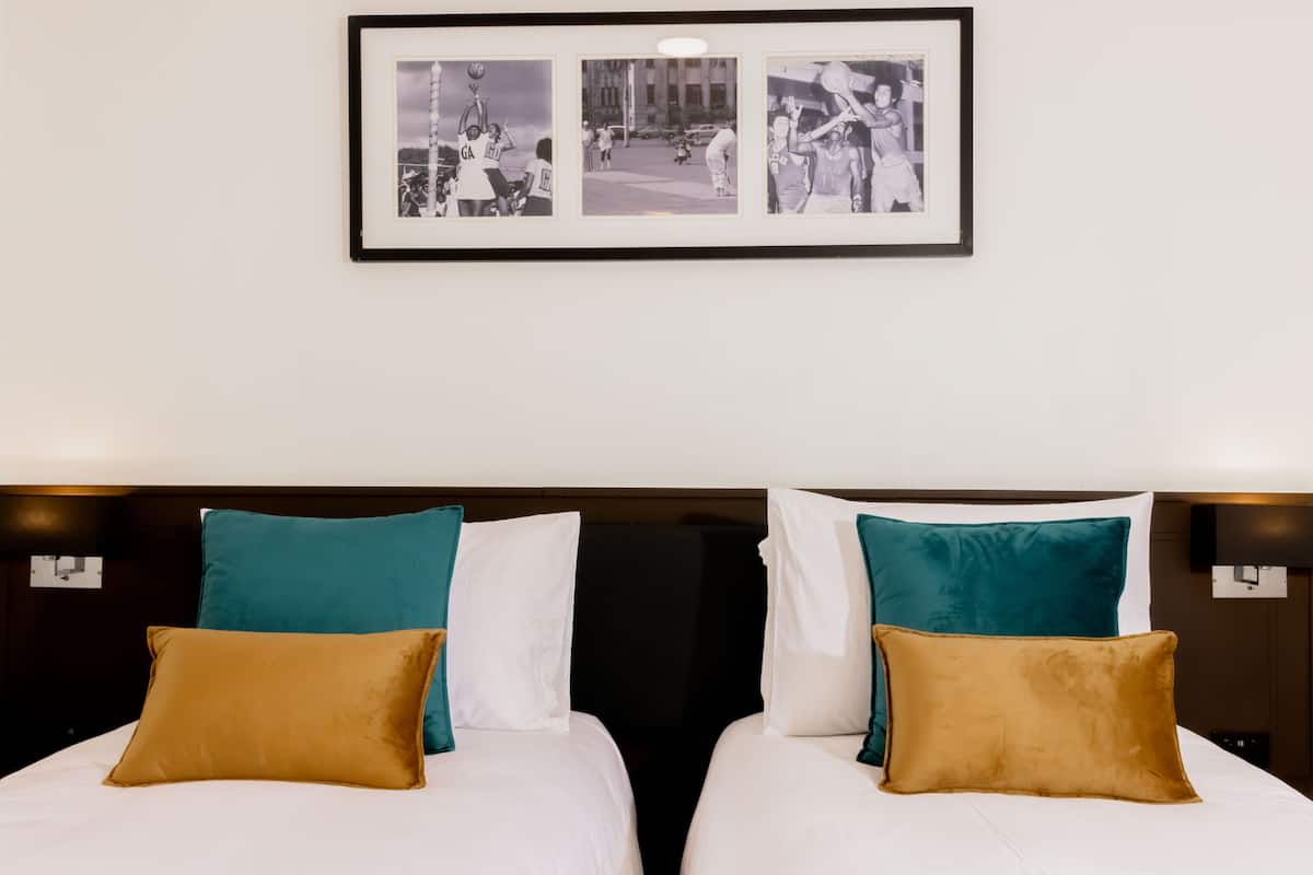 Habitación de hotel con dos camas, cada una con almohadas verde azulado y marrones, y un collage de fotos enmarcado en la pared.