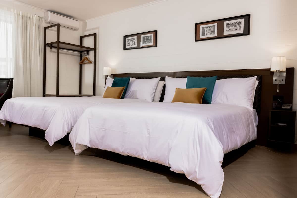 Habitación de hotel con dos camas, piso de madera, armario, escritorio y mesita de noche con lámpara.