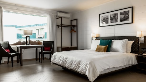 Una habitación de hotel con cama, escritorio, sillas, estante colgante y un cuadro en la pared.