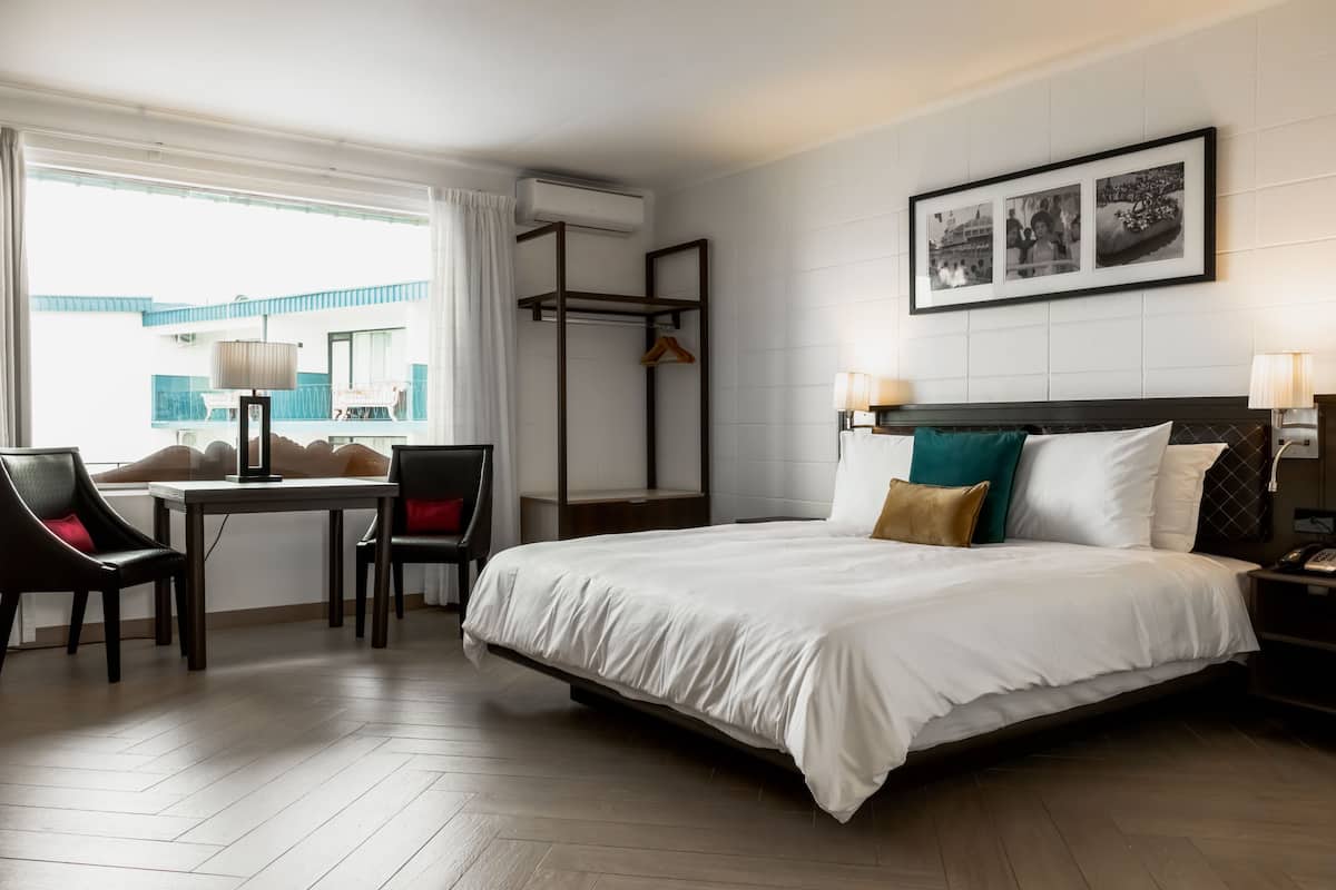 Una habitación de hotel con cama, escritorio, sillas, estante colgante y un cuadro en la pared.