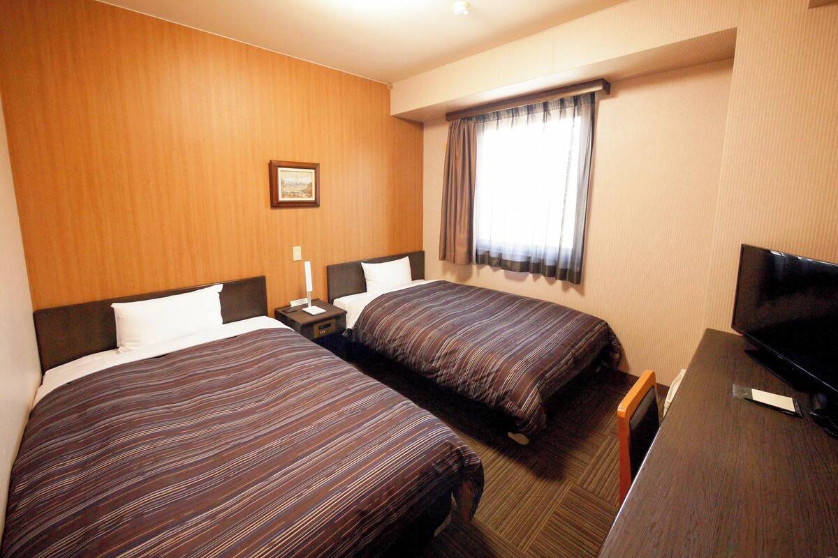 A hotel room with two beds, a TV, a desk, and a window with curtains.