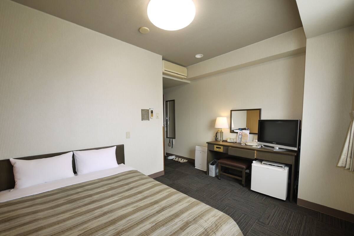 A hotel room with a bed, a desk with a television, a mirror, and a small refrigerator.