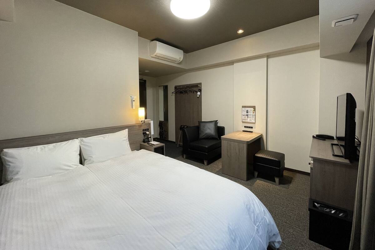 A hotel room with a large bed, a desk, a chair, a TV, and an air conditioning unit.