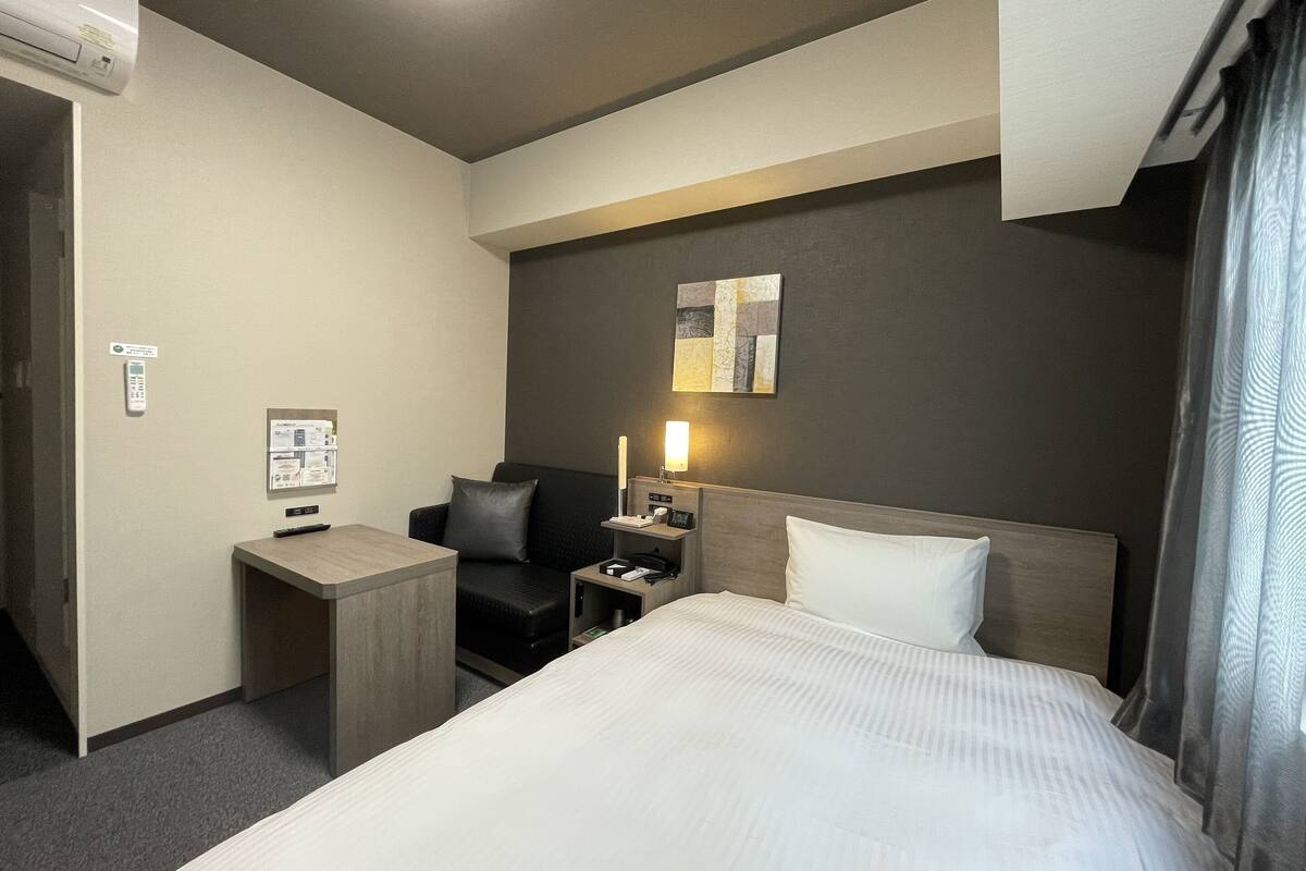 A hotel room with a bed, a desk, a chair, a nightstand, a lamp, and a wall-mounted artwork.