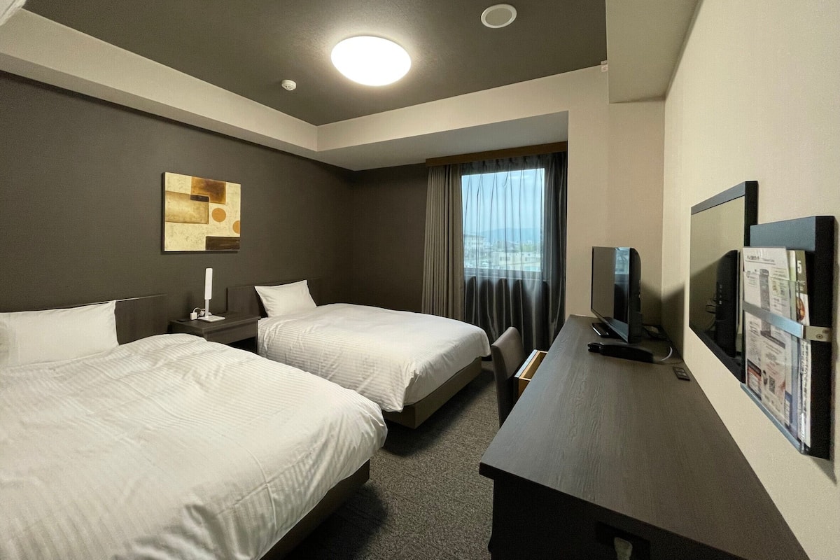 A hotel room with two beds, a desk with a television, a window with curtains, and a ceiling light.