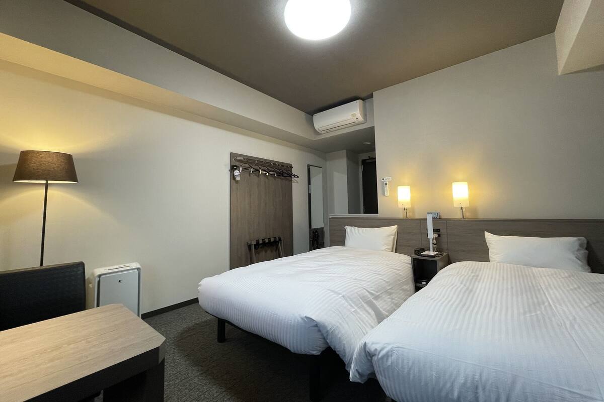 A hotel room with two beds, a desk, a lamp, and a ceiling light.
