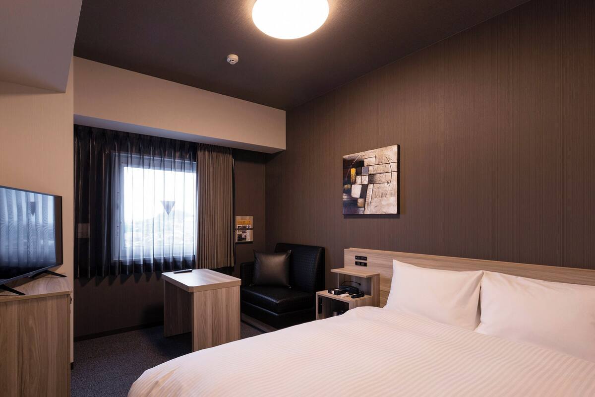 A hotel room with a large bed, a desk, a chair, a TV, and a window with curtains.