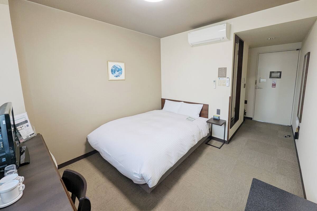 A hotel room with a bed, a desk with a computer, a chair, and a coffee table. There is a picture on the wall and an air conditioning unit above the bed.