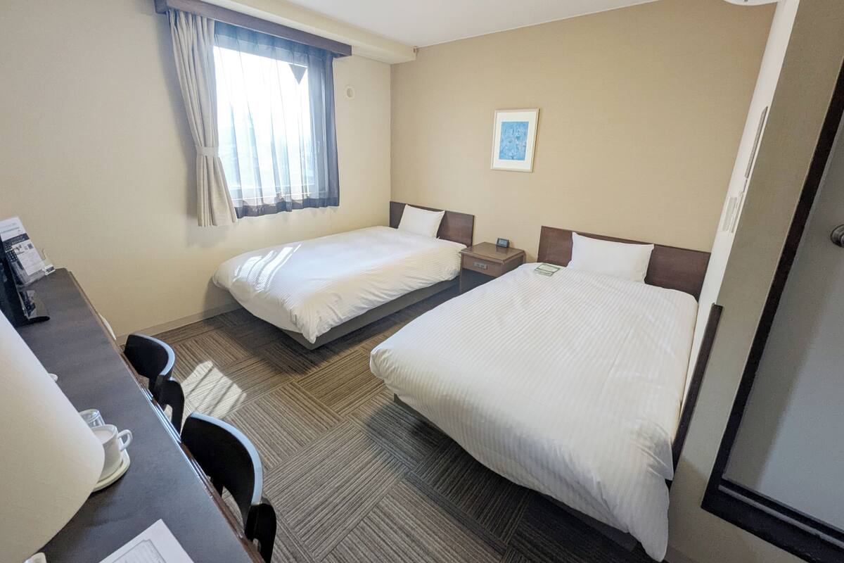 A hotel room with two single beds, a desk with a chair, a small table, and a window with curtains.