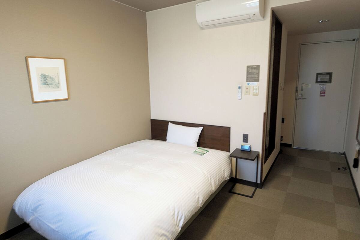 A hotel room with a single bed, a nightstand, an air conditioning unit, a framed picture on the wall, and a door leading to another room.