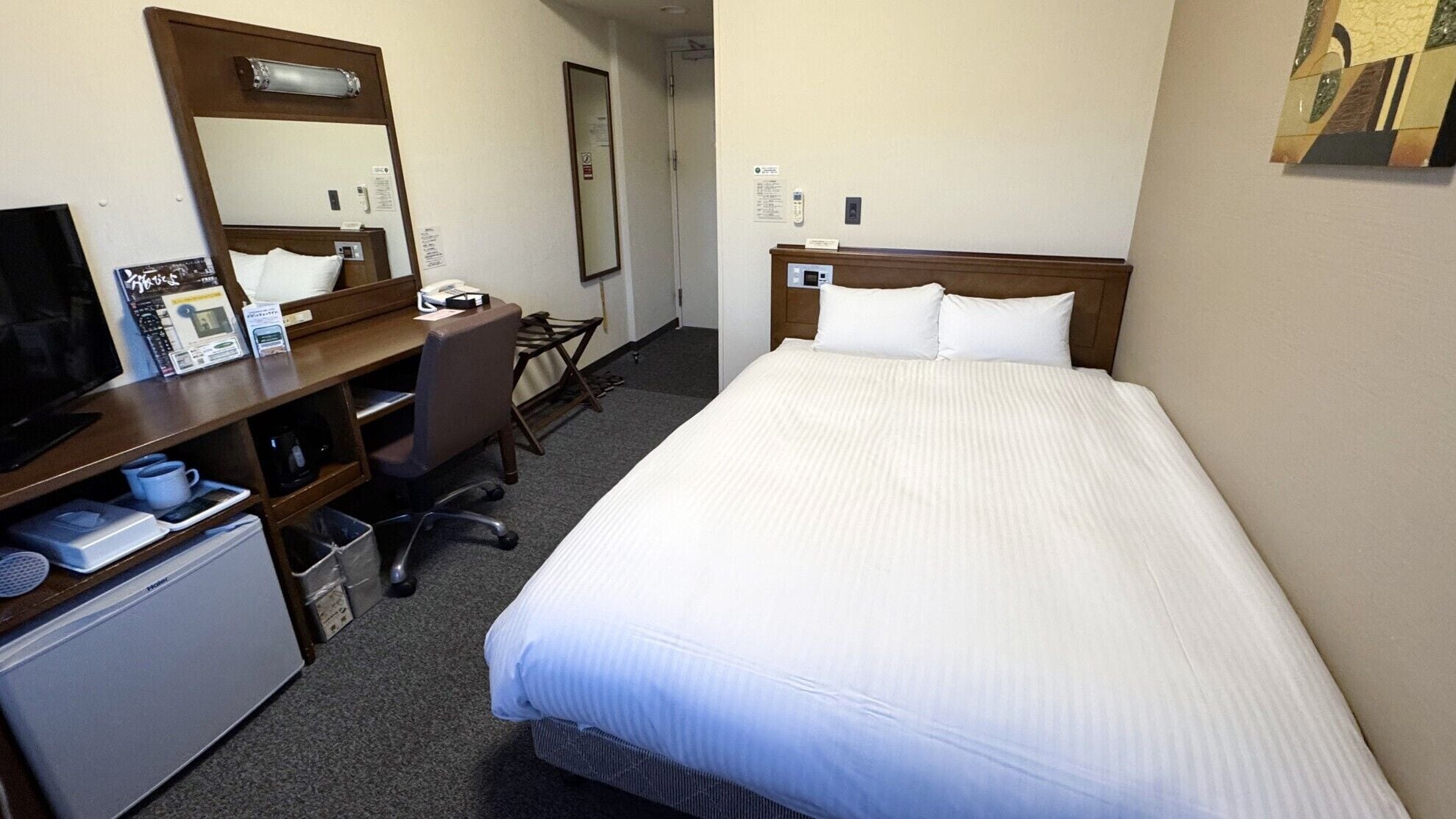 A hotel room with a bed, a desk with a computer, a chair, a small refrigerator, and a mirror.