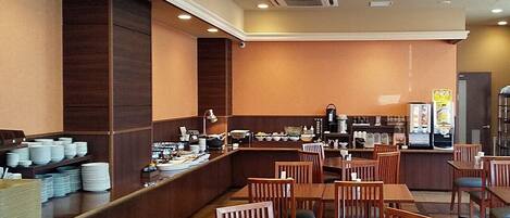 Un restaurante con mesas y sillas de madera, una barra de buffet con platos y tazones, y una máquina de café.