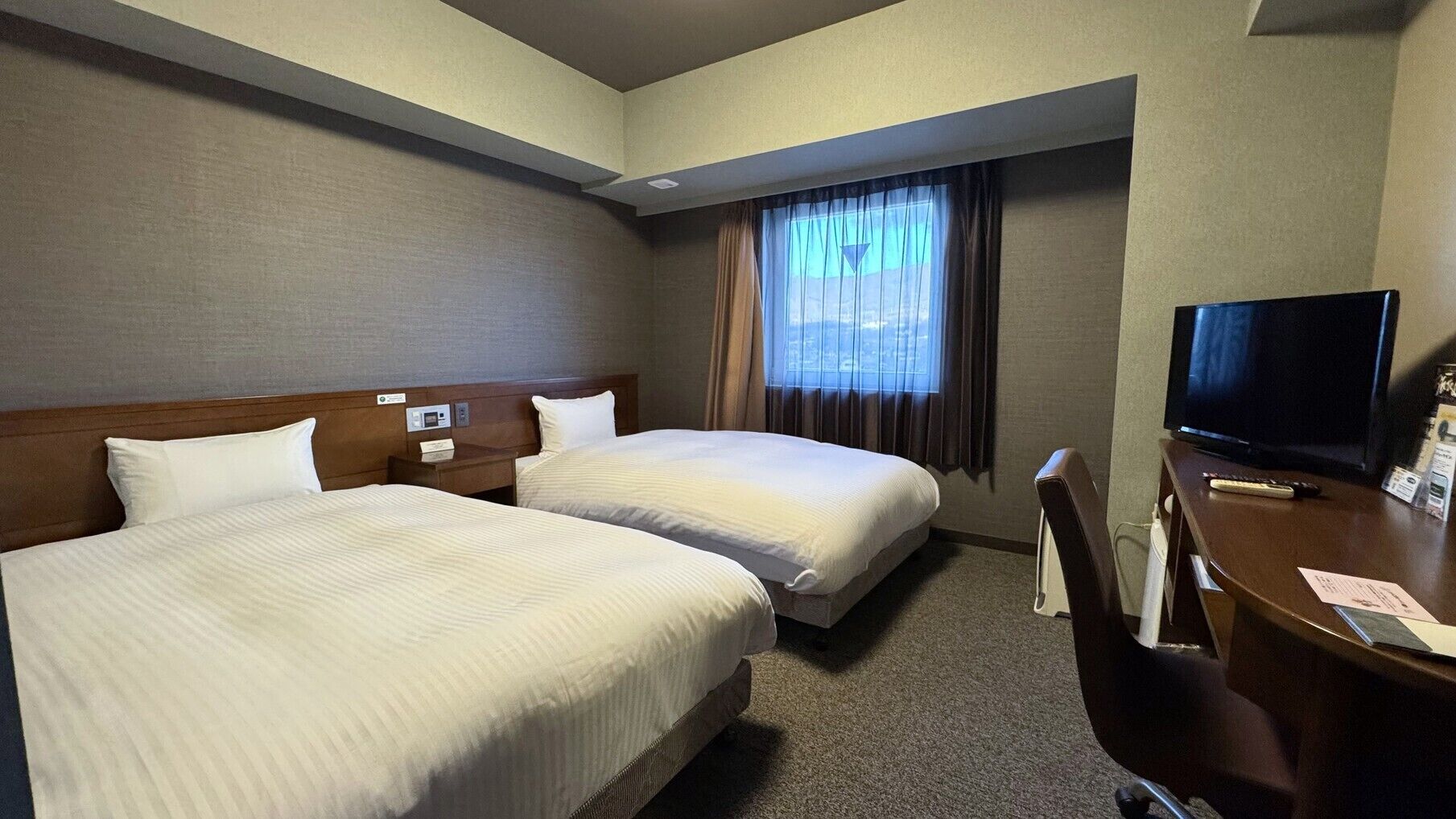 A hotel room with two beds, a desk with a TV, and a window with curtains.