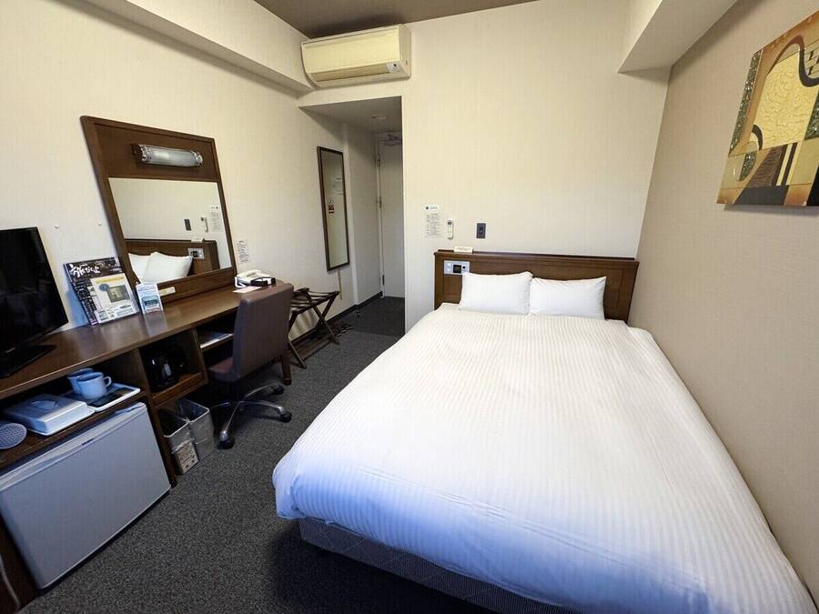 A hotel room with a bed, a desk with a computer, a chair, a small fridge, a mirror, and a painting on the wall.