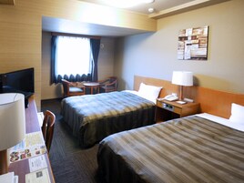 Quarto de hotel com duas camas, uma escrivaninha com computador, uma cadeira, uma mesinha e uma janela com cortinas.
