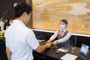 Uma mesa de recepção com divisória de vidro, uma obra de arte emoldurada representando uma pagoda e uma pequena mesa com um calendário e um telefone.