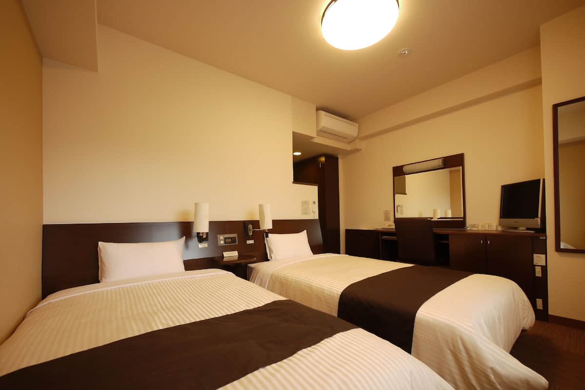 A hotel room with two beds, a desk, a chair, a mirror, and an air conditioning unit.