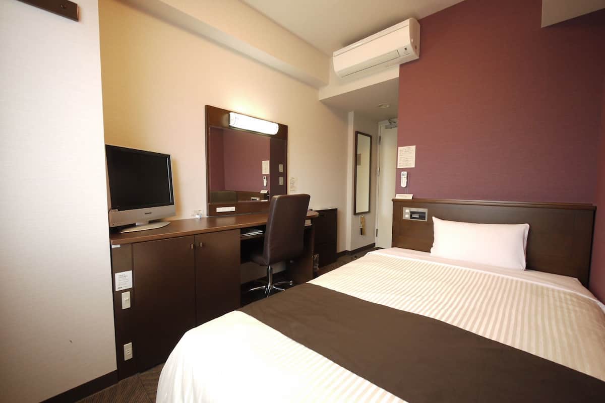 A hotel room with a bed, a desk, a chair, a television, and an air conditioning unit.