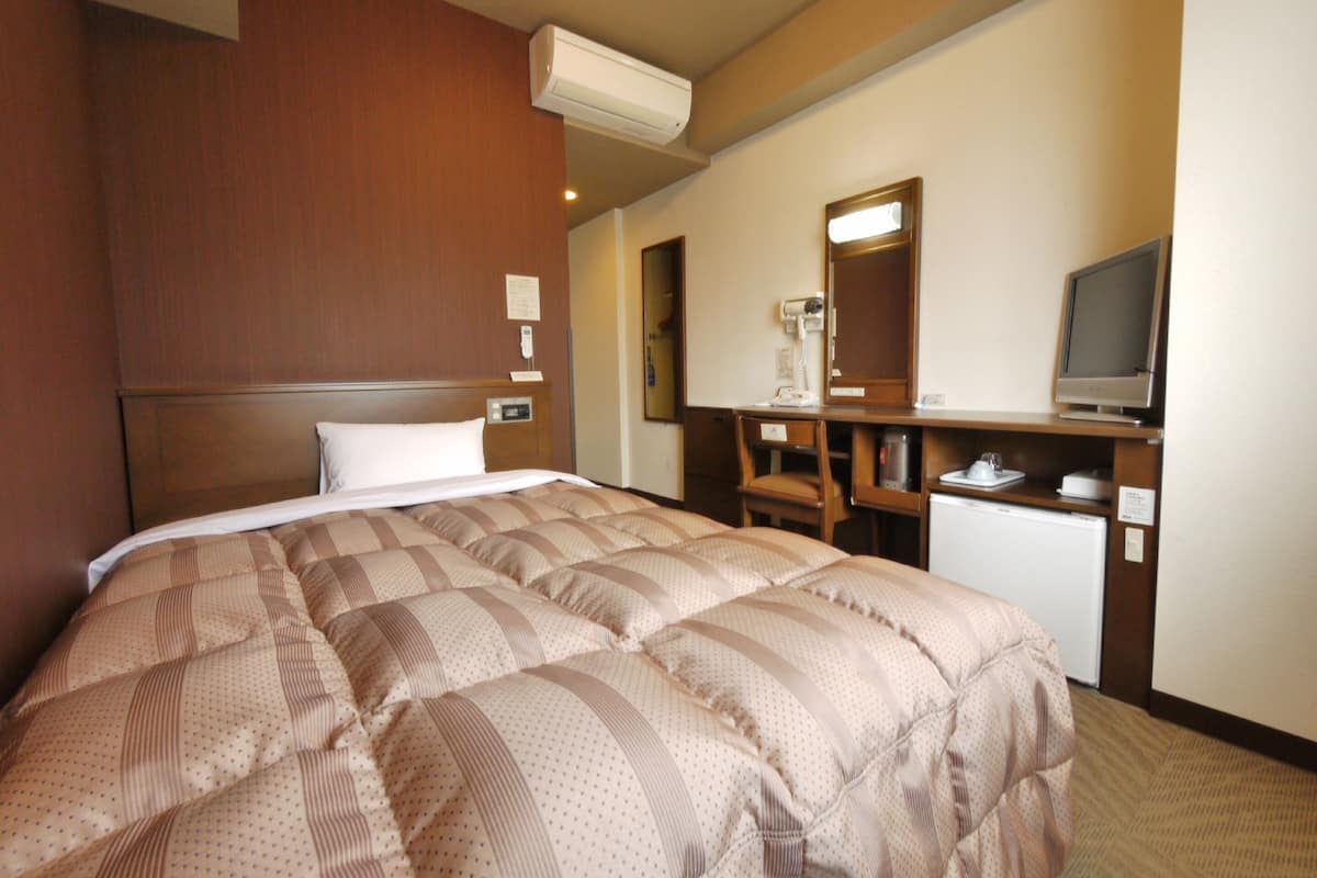 A hotel room with a large bed, a desk with a chair, a television, and an air conditioning unit.