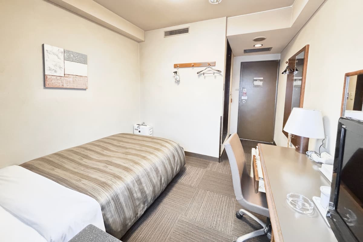 Ein Hotelzimmer mit einem Bett, einem Schreibtisch mit Stuhl, einem Fernseher, einem Spiegel und einer Tür zu einem weiteren Raum.