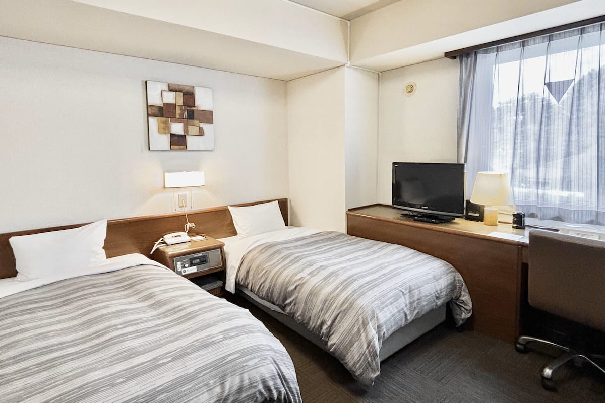 Ein Hotelzimmer mit zwei Betten, einem Schreibtisch, einem Fernseher, einer Lampe und einem fenster mit Vorhängen.