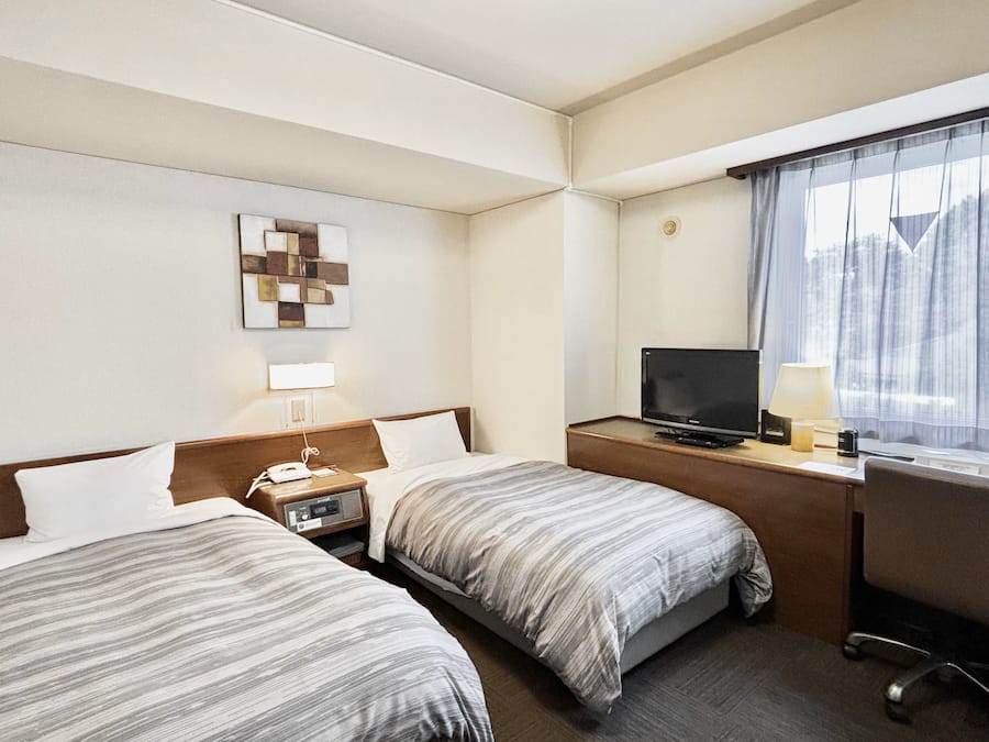Quarto de hotel com duas camas, uma mesa de trabalho com televisão, uma lâmpada e uma janela com cortinas.