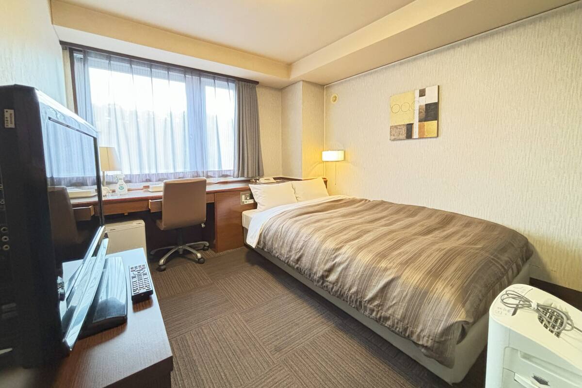 Ein Hotelzimmer mit Bett, Schreibtisch, Computer und einem fensterbepflanzten Fenster.