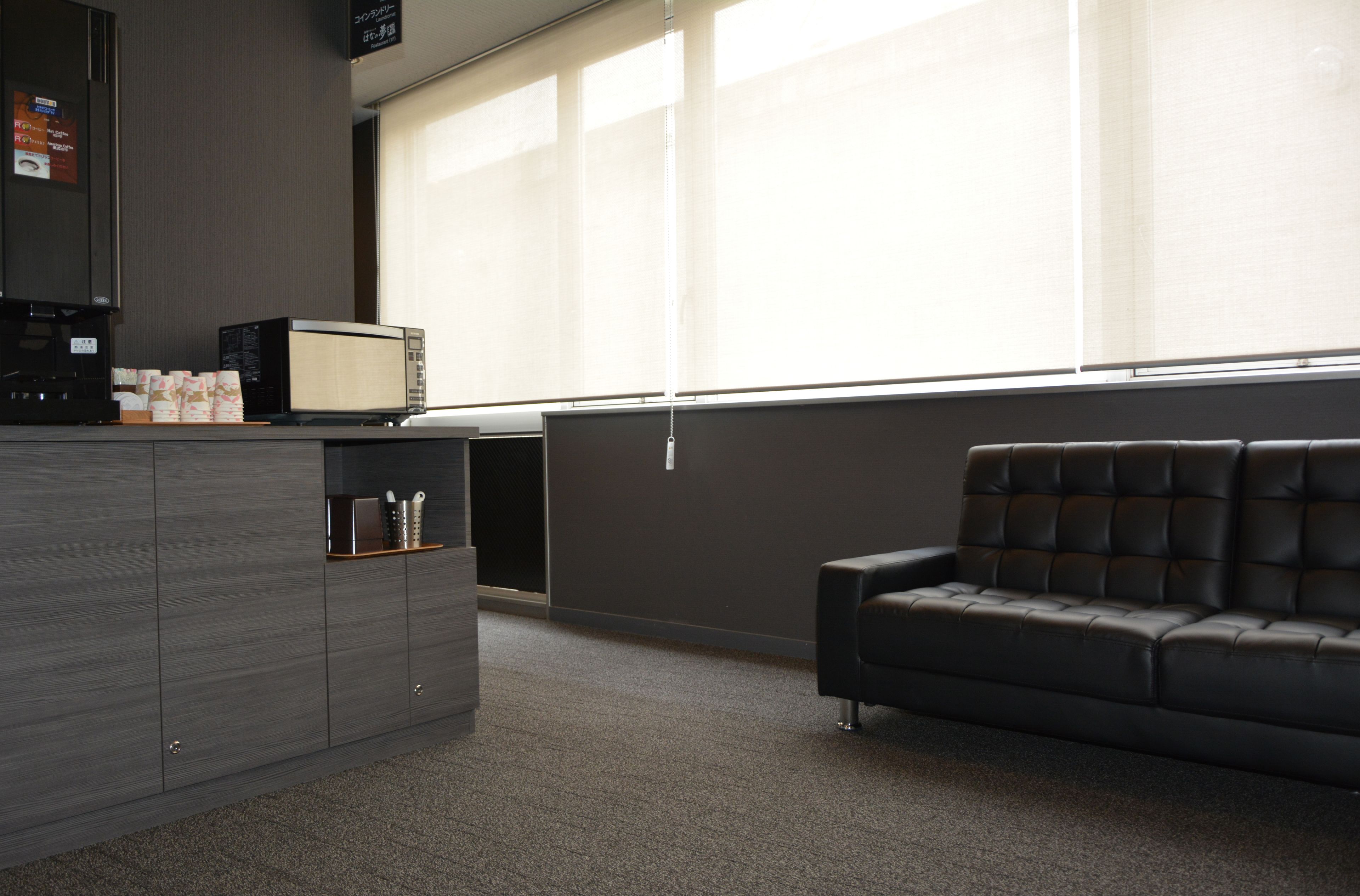 A modern office space with a dark grey cabinet, a black leather sofa, and a large window with blinds.