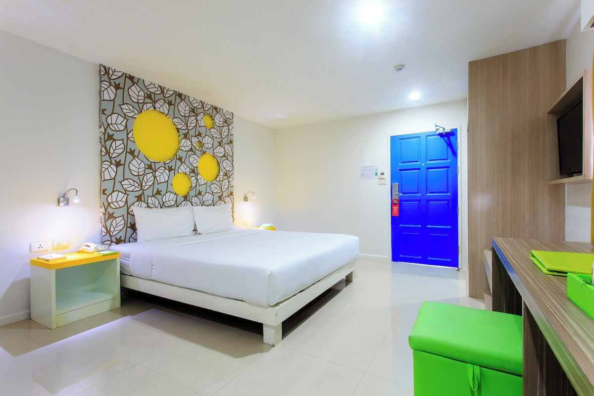 A hotel room with a bed, a nightstand, a green bench, a blue door, and a wall with a leaf pattern.