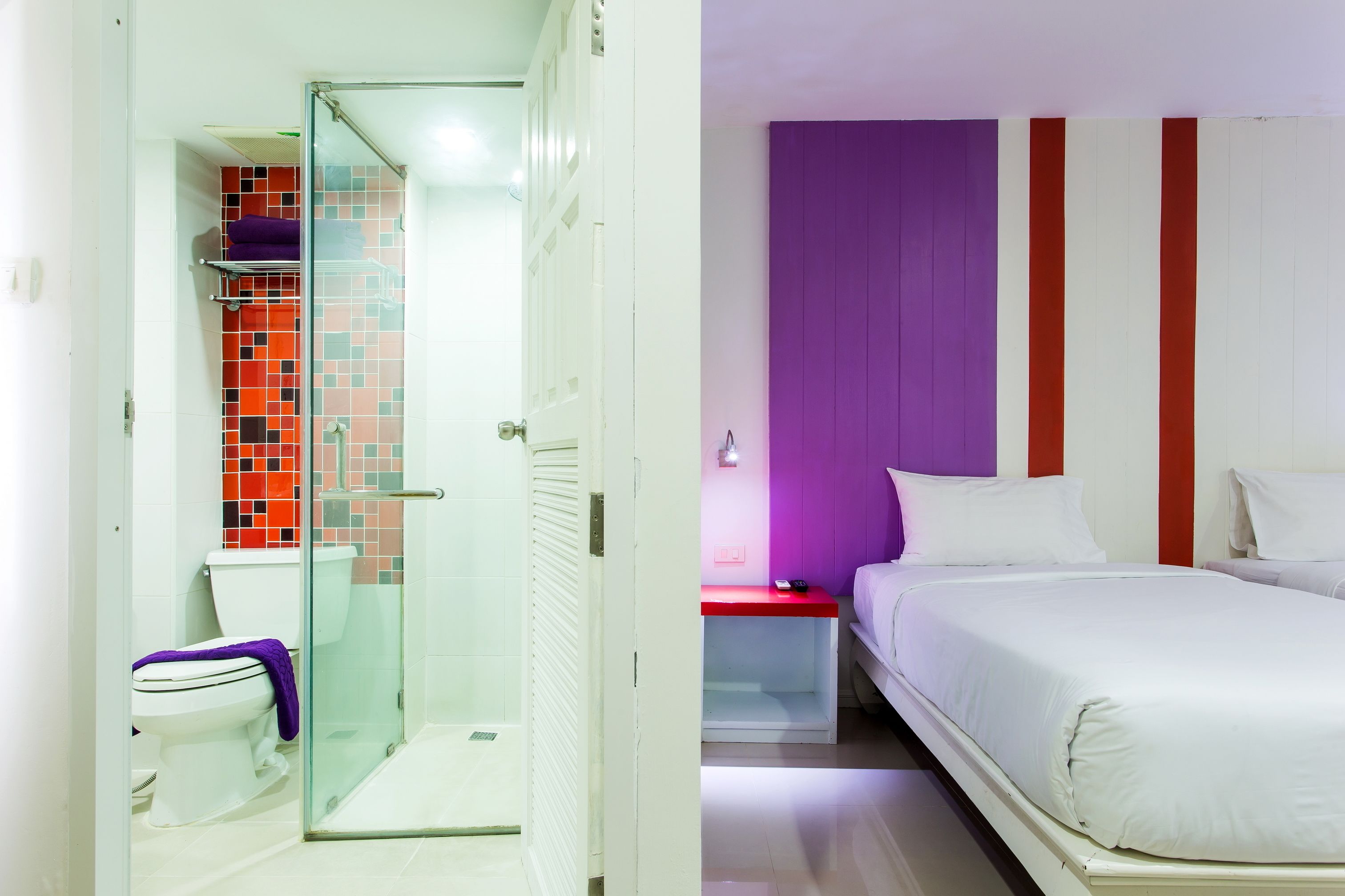A hotel room with a glass shower enclosure, a white toilet, a bed with white bedding, and a red nightstand.