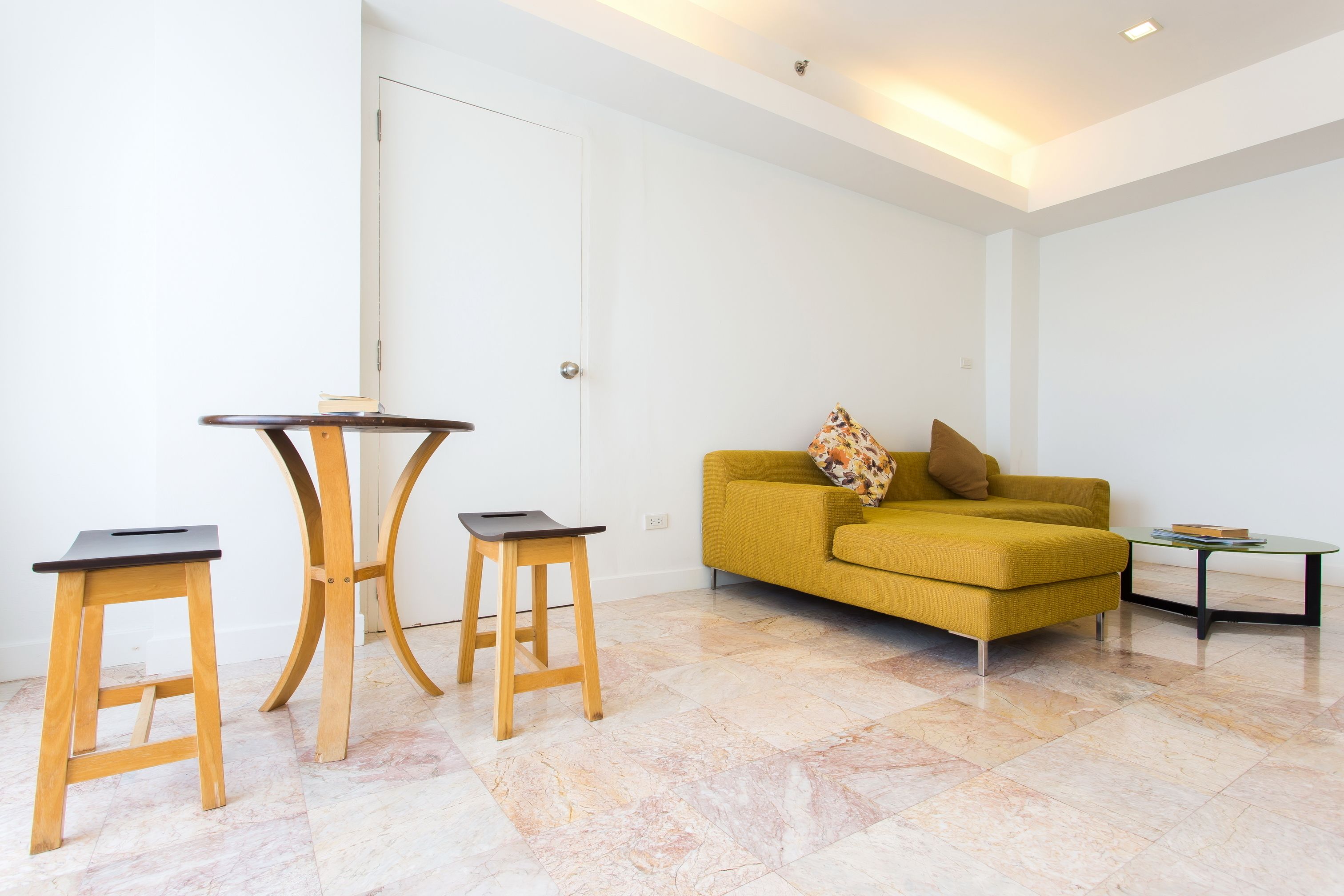 A modern living room with a yellow sofa, two wooden stools, a small round table, and a glass coffee table.