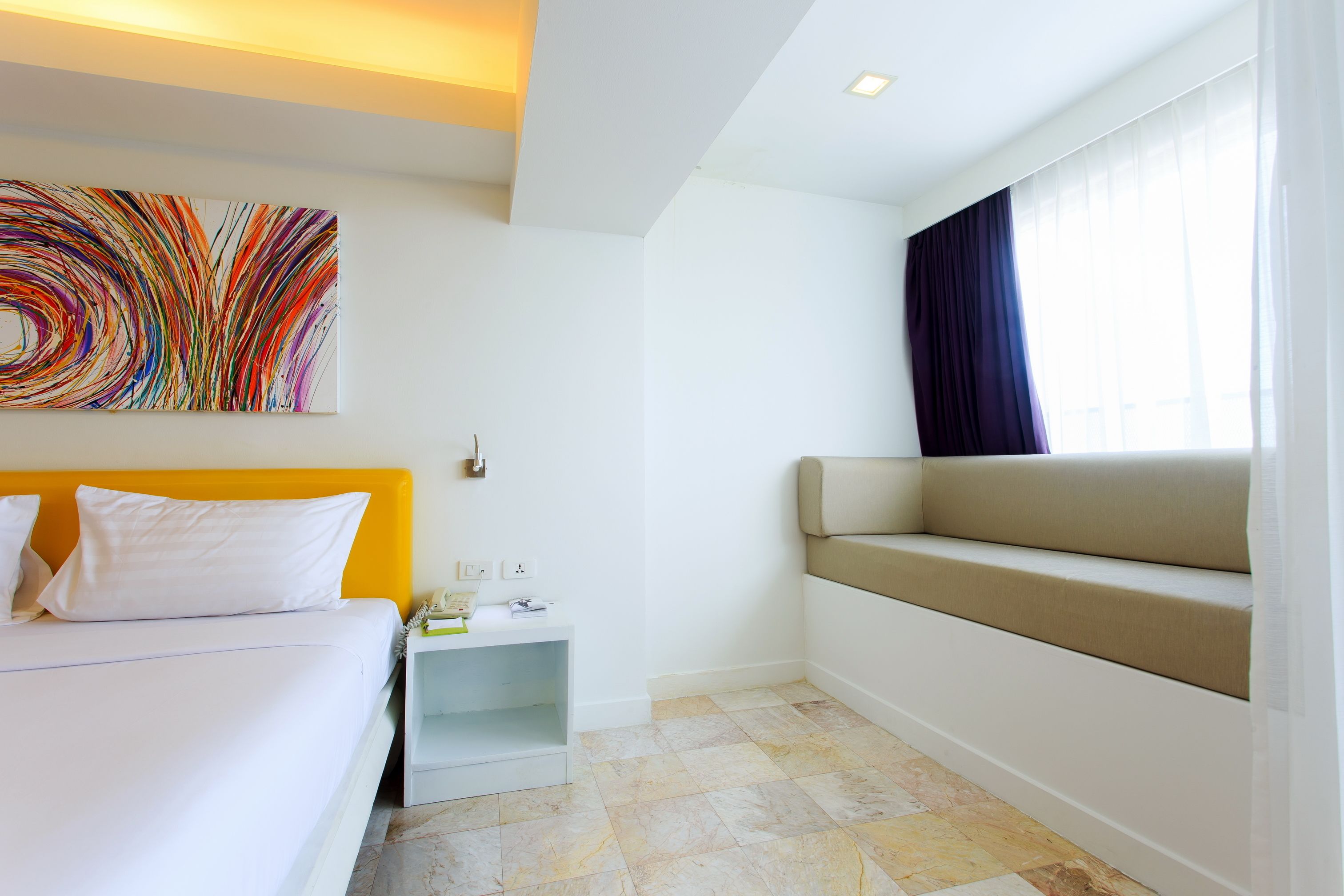 A hotel room with a bed, a bench, a colorful abstract painting, and a window with curtains.