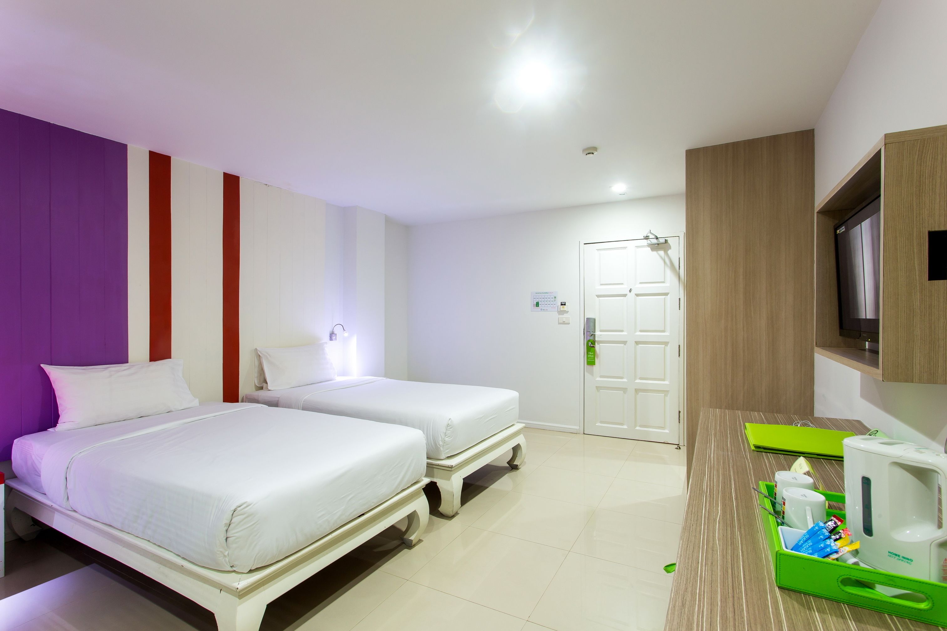 A hotel room with two beds, a wooden desk with a green tray, a white kettle, and a television on the wall.