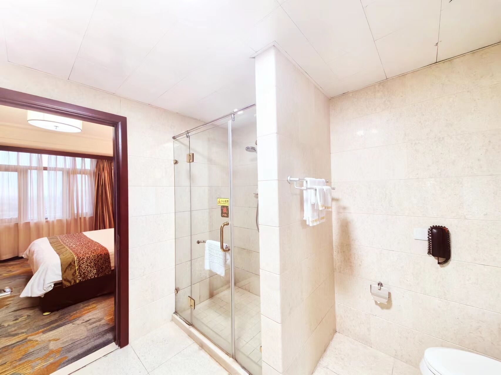 Habitación de hotel con ducha de vidrio, cama con colcha estampada y baño visible con inodoro y perchero para toallas.