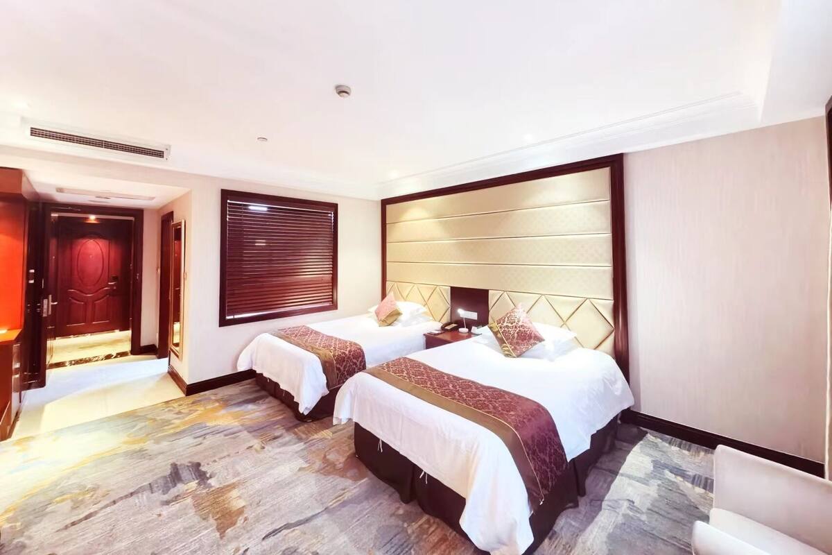 Een hotelkamer met twee bedden, een groot raam met zonwering, een hoofdeinde met een geometrisch patroon en een vloerbedekking.