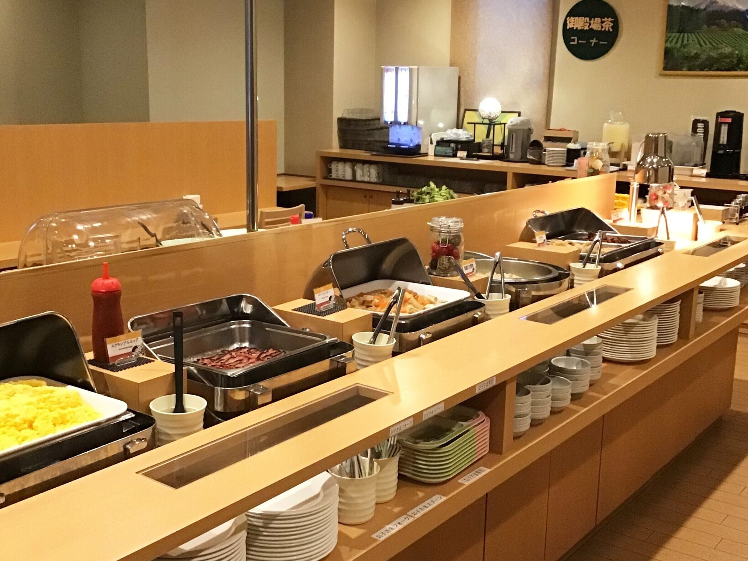A buffet setup with various food stations, including a hot sauce dispenser, a coffee machine, and a refrigerator.