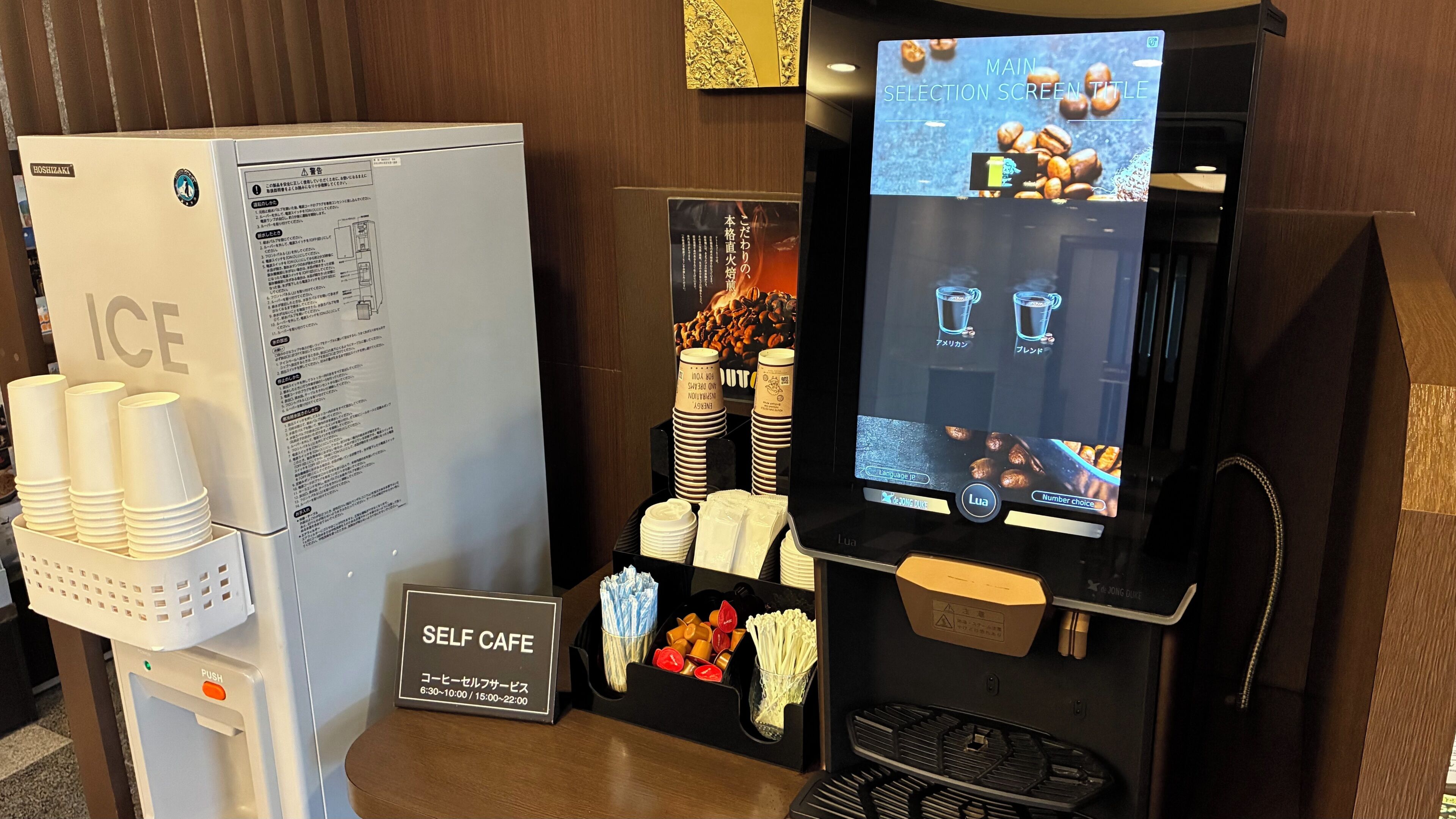 Una estación de café autoservicio con una pantalla digital que muestra el menú, un refrigerador con la etiqueta 'HIELO' y un cartel de autoservicio.