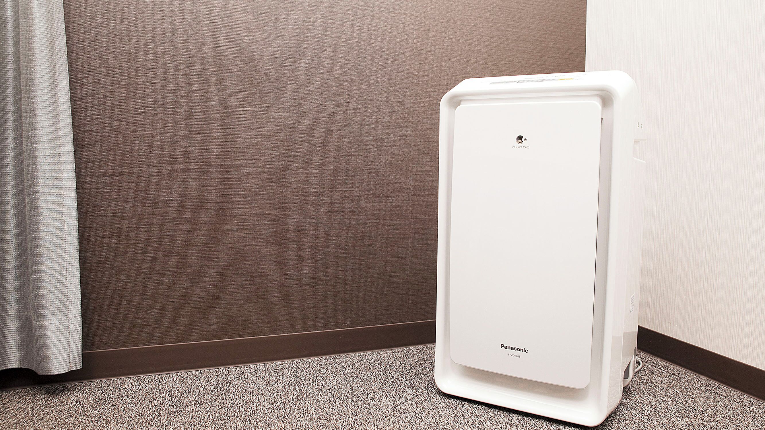 Un refrigerador blanco Panasonic en una esquina de la habitación de hotel, con una pared texturizada marrón y una cortina gris.