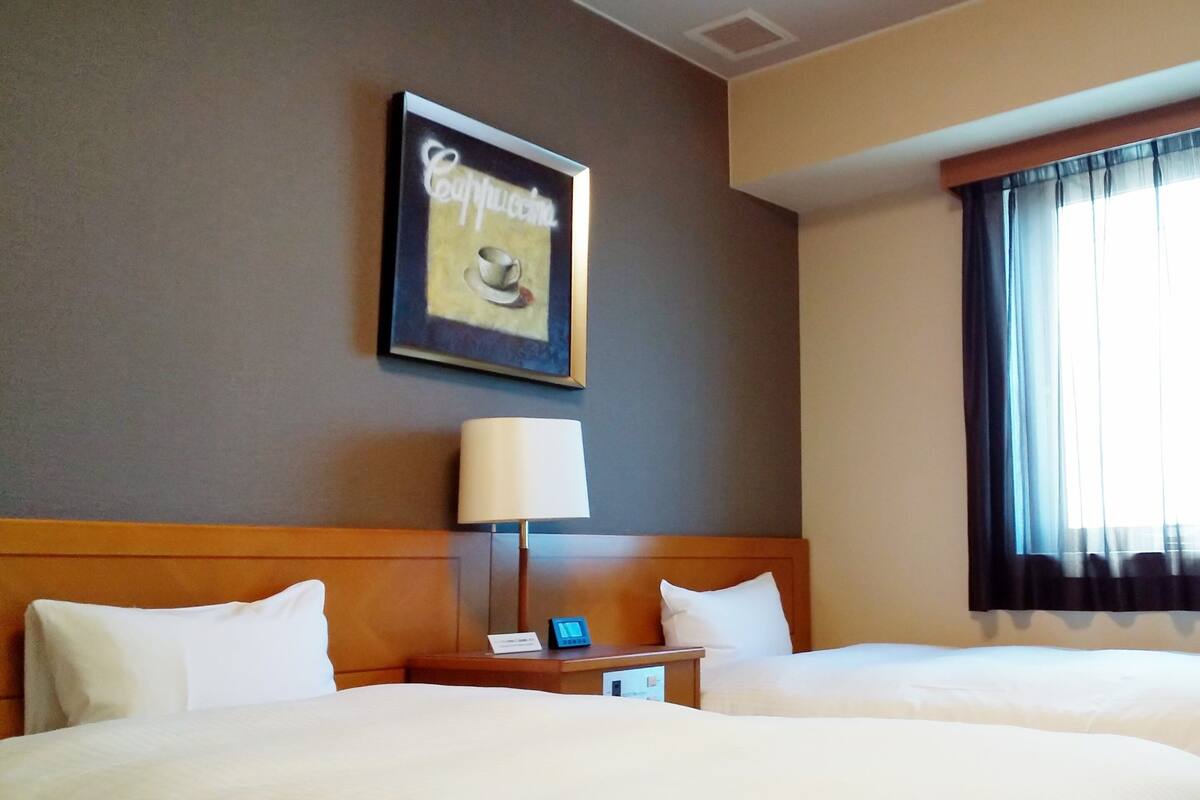 Ein Hotelzimmer mit zwei Betten, einem Nachttisch mit Lampe, einem gerahmten Bild an der Wand und einem Fenster mit Vorhängen.