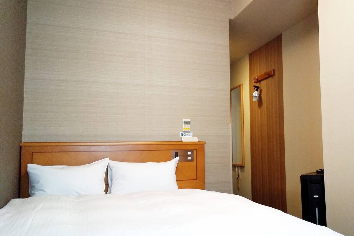 Ein Hotelzimmer mit Holz-Kopfteil, einem Bett mit weißer Bettwäsche, einem Nachttisch mit Lampe und einer Tür mit Spiegel.