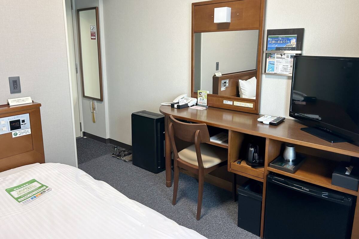A hotel room with a bed, a desk with a chair, a television, a small refrigerator, and a bedside table.