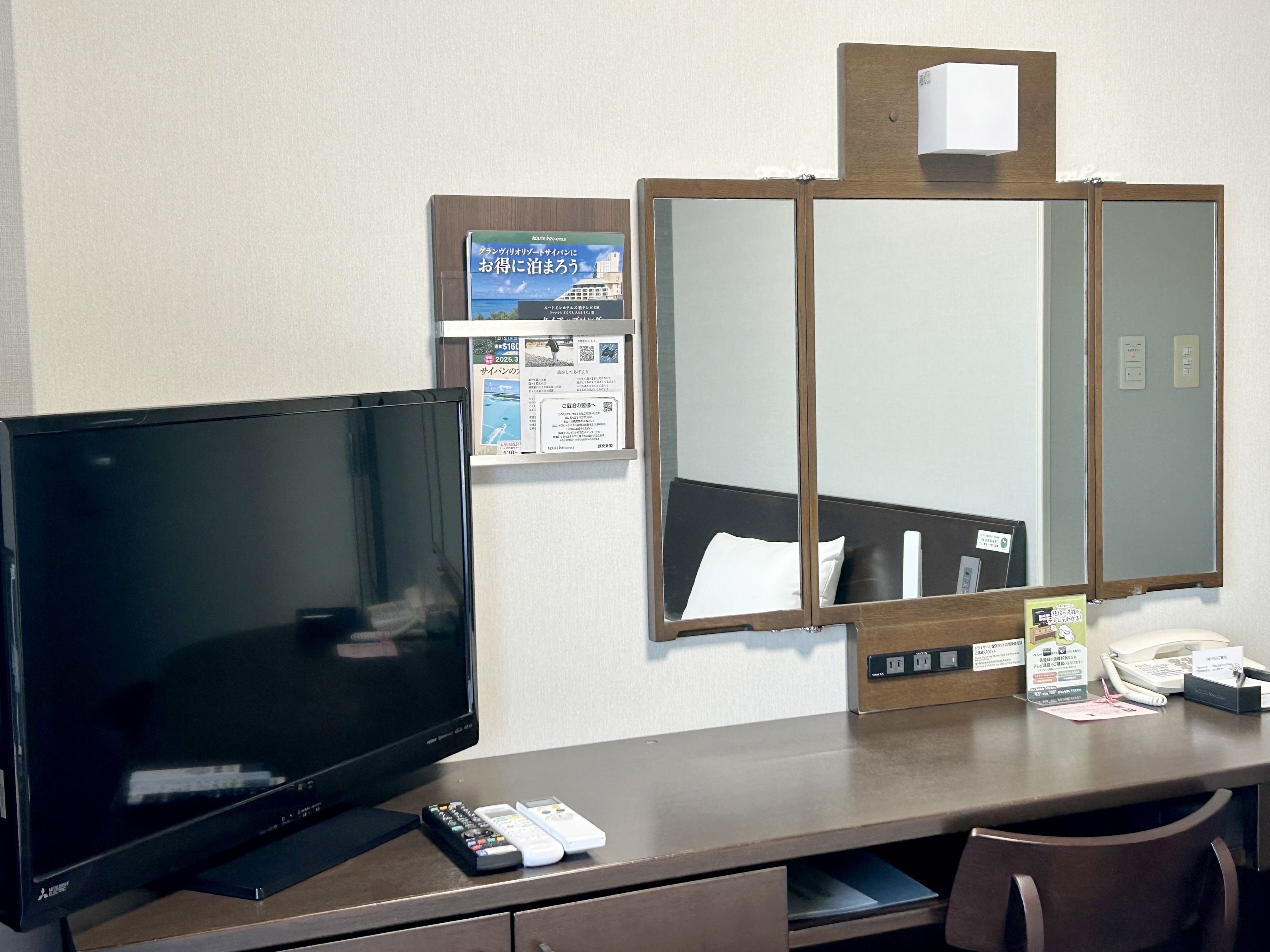 A hotel room with a television, a desk with a phone, and a mirror.