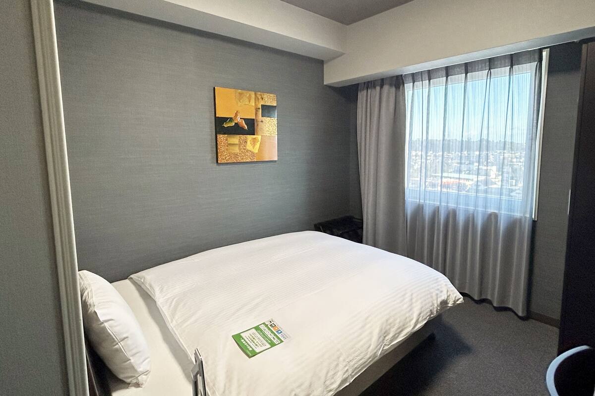 A hotel room with a bed, a window with curtains, a wall-mounted artwork, and a small table.
