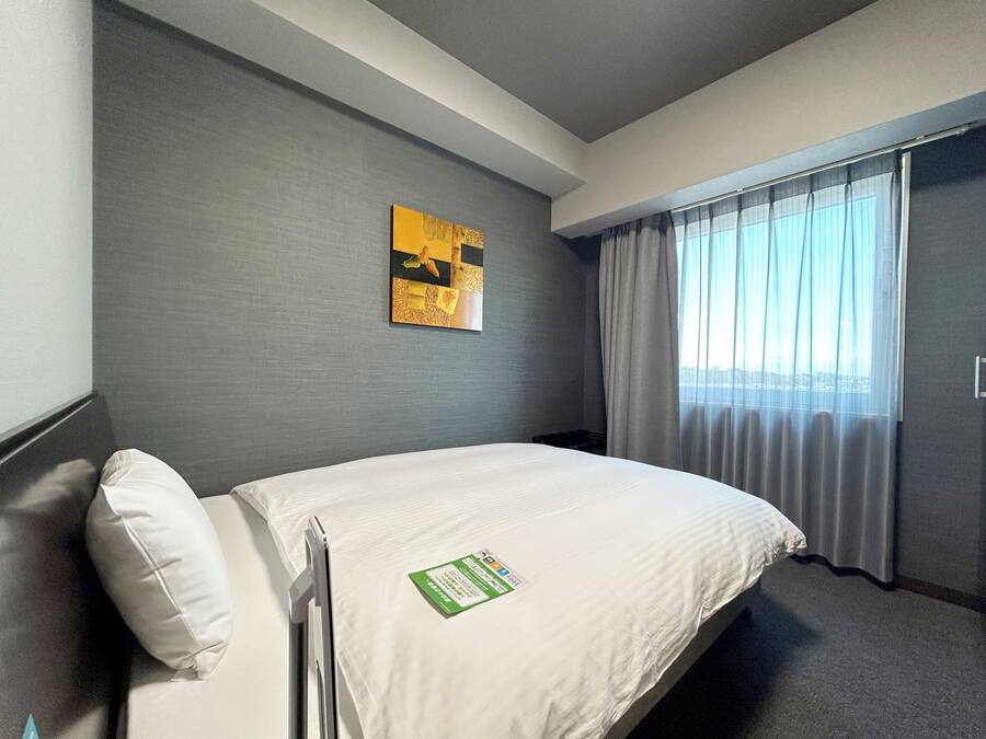 Quarto de hotel com cama, travesseiro, obra de arte na parede, janela com cortinas e porta.